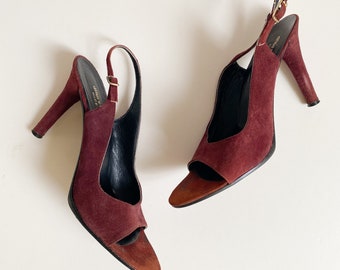 Puede incluir: Un par de sandalias de tacón de ante burdeos con dedos abiertos y tiras de slingback. Las sandalias tienen una hebilla dorada en la tira.