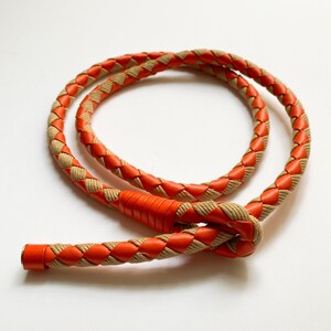 May include: A braided leather cord with a tan and orange color scheme. The cord is knotted at one end.
