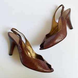 Puede incluir: Un par de zapatos de tacón slingback de cuero marrón con diseño de punta abierta. Los zapatos tienen un patrón texturizado, similar a la piel de reptil, y un tacón alto. El interior es dorado y los tacones son marrones oscuros.
