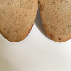 May include: Close-up of the soles of two brown leather shoes. The soles are worn and have the numbers "41" and "42" written on them in red ink.