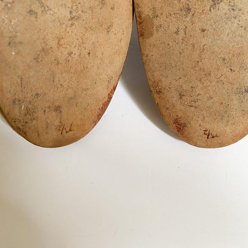 May include: Close-up of the soles of two brown leather shoes. The soles are worn and have the numbers "41" and "42" written on them in red ink.