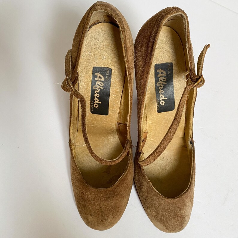 May include: A pair of brown suede heeled shoes with ankle straps. The shoes have a label on the inside that reads "This is Alfredo Greece".