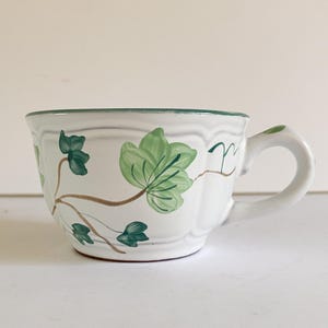 May include: White ceramic teacup with a green rim and handle. The cup is decorated with green ivy leaves and brown stems. The cup has a wide, shallow shape.