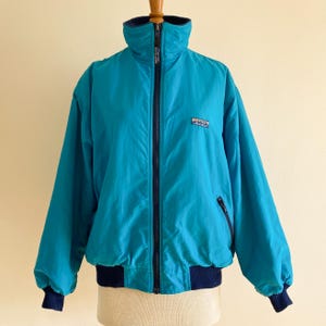May include: A teal blue vintage bomber jacket with a zipper closure and a navy blue ribbed collar, cuffs, and waistband. The jacket has a small logo patch on the left chest.