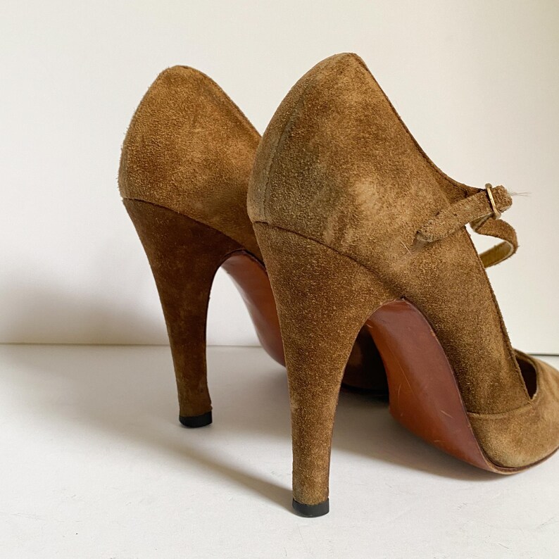 May include: A pair of brown suede high-heeled shoes with a strap and buckle. The shoes have a pointed toe and a stiletto heel.