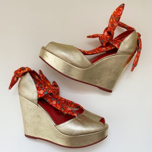 May include: A pair of gold platform wedge sandals with red and white floral fabric straps. The straps are tied around the ankles and have a bow at the back.