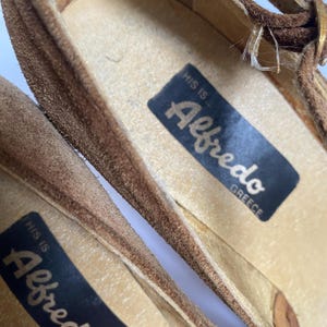 May include: Close-up of the inside of two brown suede sandals with a black label that reads "This is Alfredo Greece".