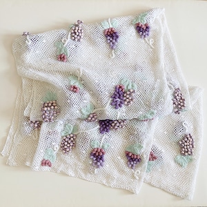 May include: White, open-weave fabric scarf or shawl, adorned with embroidered clusters of purple grapes, green leaves, and pink flowers. The grapes have a textured, beaded appearance. The design is repeated across the fabric.
