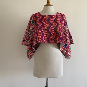 May include: A red, square-neck crop top with short sleeves. The top features a geometric pattern in blue, green, yellow, and white. The sleeves have a detailed, colorful design. The garment is displayed on a mannequin.