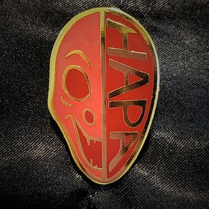 Puede incluir: Un pin rojo y dorado con la palabra "HAPA" en el lado derecho y una cara estilizada en el lado izquierdo. El pin tiene un borde dorado y está sobre un fondo negro.