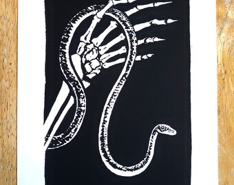 May include: Black and white art print featuring a skeletal hand holding a snake. The snake is white with a detailed pattern, and the background is solid black. The artist's signature is in the bottom right corner.