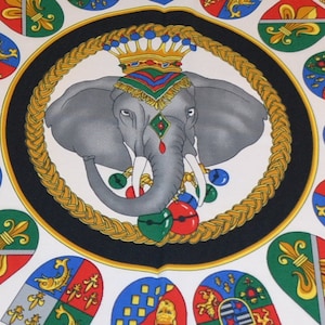 May include: A white background with a black circle in the center. Inside the circle is a gray elephant wearing a gold crown and colorful jewels. The elephant is surrounded by a gold rope design. Around the circle are 12 colorful shields with different designs.