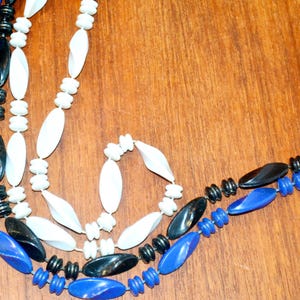 May include: Three strands of beads, one white, one black, and one blue, are intertwined. The beads are oval-shaped and have a glossy finish.