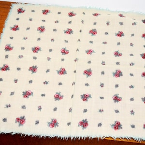 May include: Square cream textile featuring a pattern of red roses and small grey floral accents. The edges are trimmed with a light blue fringe. This textile could be a decorative item or a vintage piece.