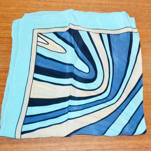 Könnte beinhalten: Ein gefalteter, quadratischer Schal mit hellblauem Rand und einem zentralen Design aus wirbelnden blauen, marineblauen und cremefarbenen Linien. Der Schal ist aus leichtem Stoff gefertigt. Der Schal liegt auf einer Holzoberfläche.