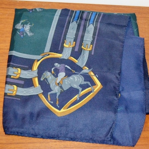May include: A square silk scarf featuring a deep navy blue and dark green background. The design includes a central gold shield emblem depicting a polo player on a horse, surrounded by light grey belt straps with gold buckles and blue stripes.