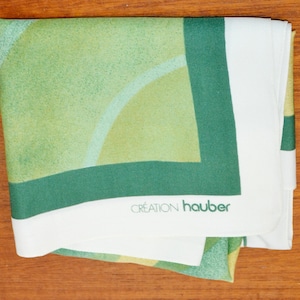 May include: A folded square scarf featuring a geometric pattern of light green and olive green circular arcs. The scarf has a wide white border and a dark green frame. The text CRÉATION hauber is printed in dark green on the white border.