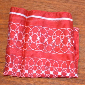 Könnte beinhalten: Ein rotes Tuch mit einem weißen geometrischen Muster aus sich überlappenden Kreisen und einem weißen Rand. Das Tuch ist gefaltet und scheint aus einem leichten Stoff zu bestehen, möglicherweise Seide oder einem ähnlichen Material.
