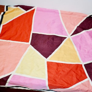 Könnte beinhalten: Ein farbenfrohes Seidentuch mit geometrischem Design. Es zeigt ein Patchwork aus Formen in Rosa-, Orange-, Gelb- und Burgundertönen, umrandet mit Weiß und mit schwarz-weißen Streifen eingefasst.