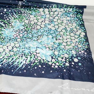 May include: A navy blue scarf with a floral pattern featuring light blue and white flowers. The scarf has a gray border and the signature "Leonardi" in the bottom right corner. The scarf is made of a soft, flowing fabric.