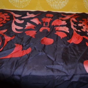 May include: A dark blue fabric featuring a repeating floral design in red hues. The fabric is draped, highlighting its texture and movement. The background is a yellow wall.