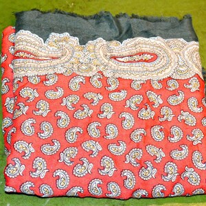 Könnte beinhalten: Gefalteter roter Paisley-Schal mit dunkelgrauem Fransen. Der Schal hat ein sich wiederholendes Paisley-Muster in Weiß und Gelb auf rotem Grund. Der obere Rand hat eine detaillierte cremefarbene und goldene Bordüre.