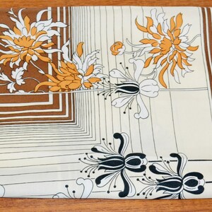 Könnte beinhalten: Ein gefalteter quadratischer Schal mit braunem Rand und floralem Design. Der Schal hat einen cremefarbenen Hintergrund mit braunen und weißen Blumenmustern und schwarzen Akzenten. Der Schal ist aus weichem, seidigem Material.