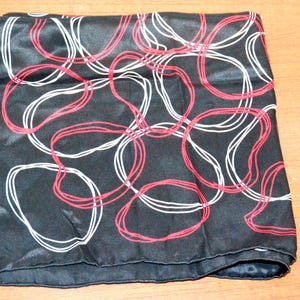 Könnte beinhalten: Schwarzes quadratisches Tuch mit einem sich wiederholenden Muster aus sich überlappenden roten und weißen Schlaufen. Das Tuch besteht aus einem glänzenden, seidigen Material und ist ordentlich gefaltet. Das Design bildet einen starken Kontrast zum schwarzen Hintergrund.
