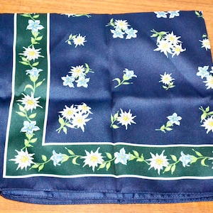 Könnte beinhalten: Ein marineblaues Seidentuch mit floralem Muster. Der Schal hat weiße und blaue Blumen, die über den Stoff verstreut sind. Ein grüner Rand mit weißen und blauen floralen Designs rahmt die Kanten ein. Das Tuch ist gefaltet und liegt auf einer Holzoberfläche.