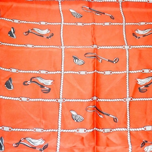 Könnte beinhalten: Ein leuchtend orangefarbener Schal mit einem sich wiederholenden Muster im Reitstil. Das Muster umfasst Reitkappen, Reitpeitschen und Pferdezubehör, alles in Schwarz und Weiß. Das Design ist auf einem Gitter aus weißem Seil angeordnet.