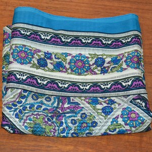 Könnte beinhalten: Gefalteter Schal mit floralem Muster in Blau-, Lila- und Grüntönen auf weißem Hintergrund. Der Schal hat einen durchgehenden türkisfarbenen Rand. Das Design umfasst stilisierte Blumen und geometrische Formen.