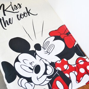 May include: A white kitchen towel featuring a black graphic of Mickey Mouse kissing Minnie Mouse. Minnie wears a red bow and a red dress with white polka dots. The text "Kiss the cook" is printed in black cursive script above the characters.