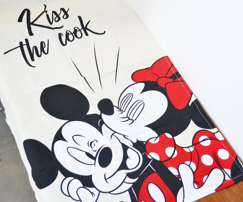 May include: A white kitchen towel featuring a black graphic of Mickey Mouse kissing Minnie Mouse. Minnie wears a red bow and a red dress with white polka dots. The text "Kiss the cook" is printed in black cursive script above the characters.
