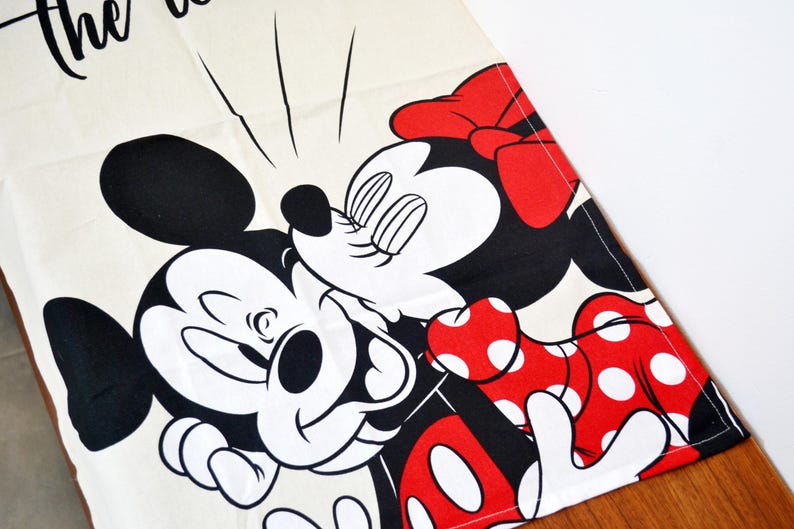 May include: A cream-colored fabric runner featuring a black and white graphic of Mickey Mouse and Minnie Mouse kissing. Minnie wears a red dress with white polka dots and a matching red bow. The text "the" is partially visible at the top edge.