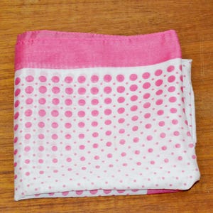 May include: A folded scarf in pink and white, featuring a polka dot design. The top edge is a solid pink, while the main body is white with pink polka dots of varying sizes. The scarf is neatly folded on a wooden surface.