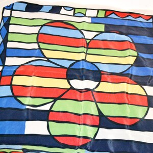 Könnte beinhalten: Ein farbenfrohes Tuch mit floralem Muster. Das Design zeigt eine große Blume mit roten, gelben, grünen und blauen Blütenblättern. Der Hintergrund hat horizontale Streifen in verschiedenen Farben und die Ränder geometrische Muster.