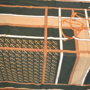 May include: A dark green scarf featuring a repeating pattern of small gold stirrups on one side and a large graphic of brown and white leather straps with buckles arranged in a grid. The design includes a prominent orange stirrup illustration.