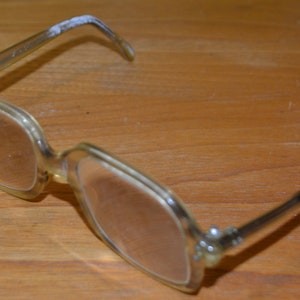 May include: A pair of clear plastic eyeglasses with square frames. The glasses are on a wooden surface.