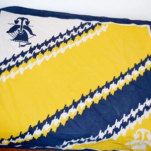 Könnte beinhalten: Ein blaues und gelbes Stoffquadrat mit weißem Rand. Der Stoff hat ein diagonales Muster aus weißem und blauem Hahnentritt. Die obere linke Ecke zeigt eine schwarze Silhouette eines Schiffes mit einem Segel.