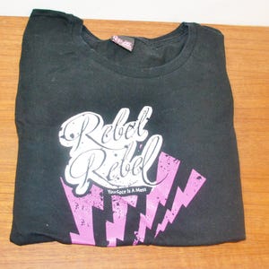 Könnte beinhalten: Schwarzes T-Shirt mit einem weißen Aufdruck, der "Rebel Rebel" mit rosa Blitzen und dem Text "Your face is a mess" lautet.