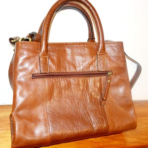 Peut inclure: Un sac à main en cuir marron avec une poignée supérieure et une bandoulière. Le sac a une poche zippée à l'avant et un tirette de fermeture éclair dorée.