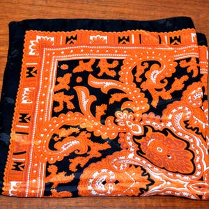 Könnte beinhalten: Ein schwarzer Seidenschal mit einem orange-weißen Paisley-Muster. Der Schal hat einen Rand aus orange-weißen geometrischen Formen.