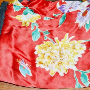 Op de afbeelding: Een rode zijden sjaal met een bloemenpatroon. De bloemen zijn geel, wit en paars. De sjaal heeft een zwarte rand.