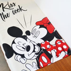 May include: A cream-colored kitchen towel featuring a black graphic of Mickey Mouse and Minnie Mouse kissing. The text "Kiss the cook" is printed in black script at the top. Minnie Mouse wears a red bow and a red dress with white polka dots.