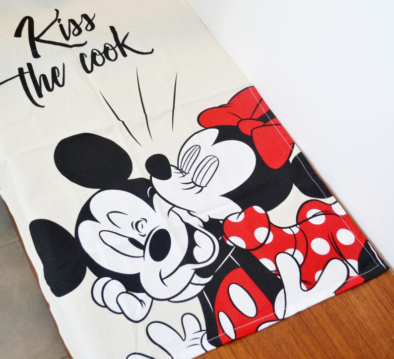 May include: A cream-colored kitchen towel featuring a black graphic of Mickey Mouse and Minnie Mouse kissing. The text "Kiss the cook" is printed in black script at the top. Minnie Mouse wears a red bow and a red dress with white polka dots.