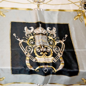 Könnte beinhalten: Seidenschal mit einem zentralen schwarzen Quadrat mit einem goldenen und silbernen Wappen, umgeben von einem grauen Rand und goldenen Akzenten. Der Schal hat einen cremefarbenen Hintergrund mit goldenen Seil- und Schnallendetails.