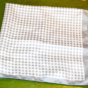 Könnte beinhalten: Ein gefaltetes, quadratisches Einstecktuch mit weißem und grauem Karomuster. Das Einstecktuch hat einen hellgrauen Rand. Der Stoff scheint Seide oder ein ähnliches leichtes Material zu sein.
