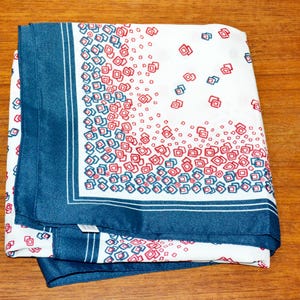 Könnte beinhalten: Ein gefalteter weißer Schal mit einem dunkelblauen Rand und einem Muster aus roten und blauen Quadraten. Der Schal ist aus einem seidigen Material gefertigt und liegt ordentlich auf einer Holzoberfläche.