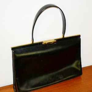 Puede incluir: Un bolso negro vintage con un asa curva y herrajes dorados. El bolso rectangular tiene un acabado brillante y un cierre de broche. El asa está sujeta a la parte superior del bolso con metal dorado. El bolso está sobre una superficie de madera.