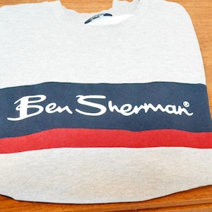 Puede incluir: Una sudadera gris doblada con una franja azul marino y roja en el pecho. Las palabras "Ben Sherman" están impresas en blanco sobre la franja azul marino. La sudadera está hecha de un material suave.